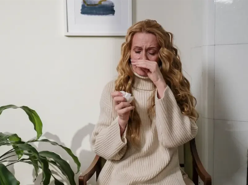 A blonde woman sitting indoors while rubbing her nose and holding a tissue due to allergy symptoms.