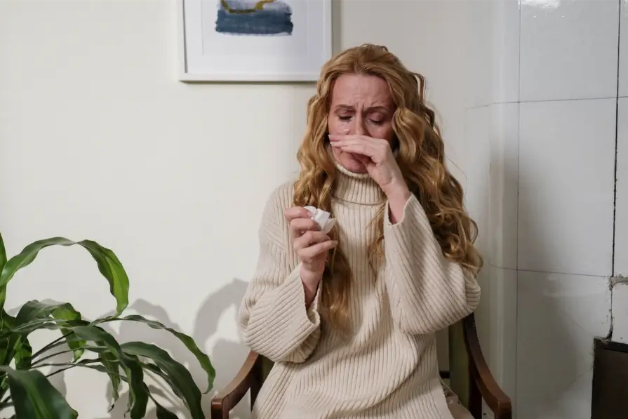 A blonde woman sitting indoors while rubbing her nose and holding a tissue due to allergy symptoms.